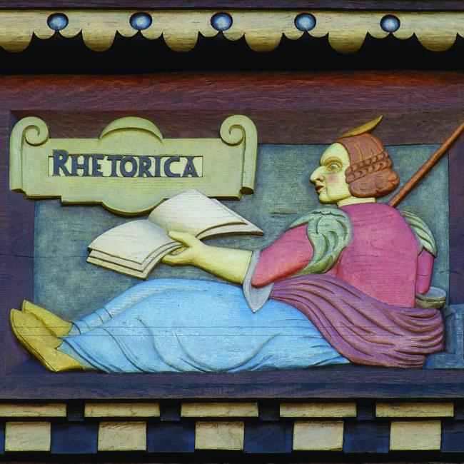 a wooden carving of a man holding a book, behind him there is a sign that says rhetorica