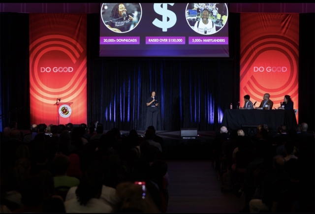 T-Kea Blackman Wide Shot Presenting at Do Good Challenge Finals; T-Kea on a dark stage with a microphone and projector in the background