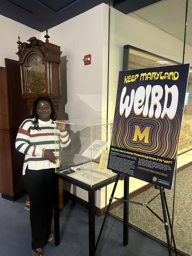 Felicity Dogbatse with "Keep Maryland Weird" poster, next to an archival display in Hornbake Library, smiling 